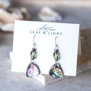 May include: Dangle earrings on a white display card labeled LEAF & LIGHT. Each earring features a silver-tone hook, a small purple bead, a translucent green glass bead, and an iridescent, multi-colored abalone shell pendant at the base.