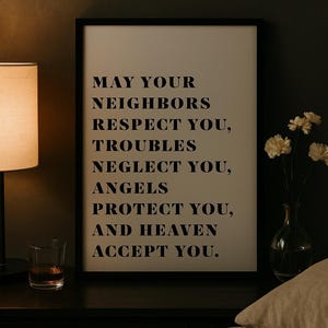Pode incluir: Uma impressão emoldurada com o texto "MAY YOUR NEIGHBORS RESPECT YOU, TROUBLES NEGLECT YOU, ANGELS PROTECT YOU, AND HEAVEN ACCEPT YOU." A impressão está em uma moldura preta, repousando sobre uma superfície escura, com uma lâmpada e um copo de líquido por perto.