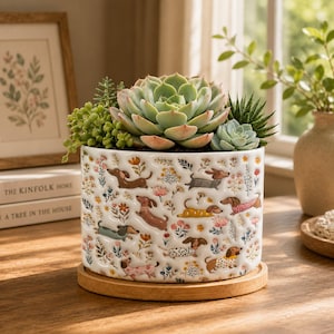 May include: A white ceramic cylindrical planter featuring a repeating pattern of colorful dachshunds and small floral illustrations. The planter sits on a circular wooden base and contains a variety of green succulents, including a large central rosette.
