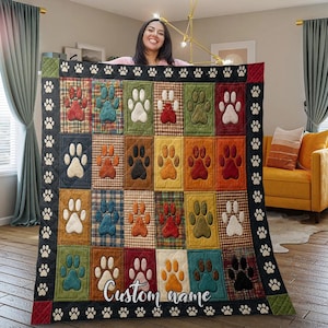 May include: A quilted throw blanket featuring a grid of rectangular panels with various plaid patterns in red, blue, green, and yellow. Each panel displays a single paw print in a contrasting color. The black border has white paw prints.