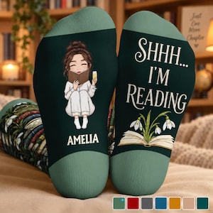 May include: A pair of dark green socks featuring a cartoon illustration of a person reading a book while holding a glass, with the name AMELIA printed below. The other sock displays the text SHHH... I'M READING above an open book with flowers.