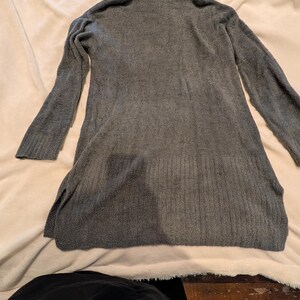 May include: A gray, long-sleeved sweater dress with a turtleneck collar. The dress has a soft, textured appearance and side slits at the hem. The garment is displayed on a light-colored surface.