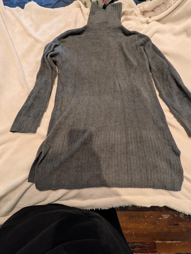 May include: A gray, long-sleeved sweater dress with a turtleneck collar. The dress has a soft, textured appearance and side slits at the hem. The garment is displayed on a light-colored surface.