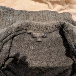 May include: A close-up of a gray, plush zip-up sweater. The sweater has a high collar and a tag that reads "Barefoot Dreams CozyChic Life". The fabric appears soft and textured, with a visible zipper.