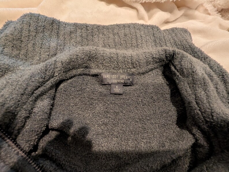 May include: A close-up of a gray, plush zip-up sweater. The sweater has a high collar and a tag that reads "Barefoot Dreams CozyChic Life". The fabric appears soft and textured, with a visible zipper.