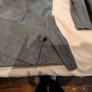 May include: A gray, textured sweater with long sleeves. The sweater is made of a soft, plush material and is lying on a cream-colored surface. The sleeves are visible, and the sweater appears to be folded.