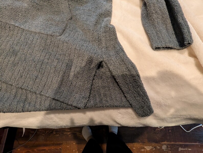 May include: A gray, textured sweater with long sleeves. The sweater is made of a soft, plush material and is lying on a cream-colored surface. The sleeves are visible, and the sweater appears to be folded.