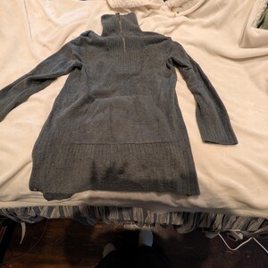 Könnte beinhalten: Ein graues Langarm-Strickkleid mit hohem Reißverschlusskragen. Das Kleid hat eine gerippte Textur und eine lockere Passform. Die Ärmel sind lang und der Saum fällt unter die Hüften. Das Kleid ist aus weichem Strickmaterial.