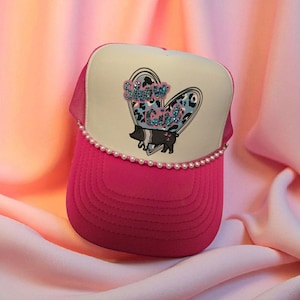 May include: A hot pink trucker hat with a white front panel featuring a heart-shaped graphic with the text "Show Girl" and a black and white pig illustration. The brim is also hot pink, and the hat is trimmed with small white pearls.