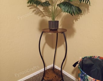 May include: A decorative plant stand with a round, wood-look top and bottom shelf. The stand has a black metal frame with curved legs. A potted plant with green leaves sits on top.