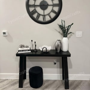 May include: A black console table with decorative items, including a large clock, vases, books, and candle holders. A black, round, pleated ottoman sits below the table. The decor is modern and minimalist.