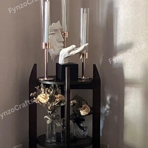 Pu&ograve; includere: Una mensola scura a tre livelli con oggetti decorativi. Presenta tre candelabri in vetro con candele bianche, una scultura bianca di un volto e una scultura di una mano bianca. I ripiani contengono fiori secchi in vasi di vetro.