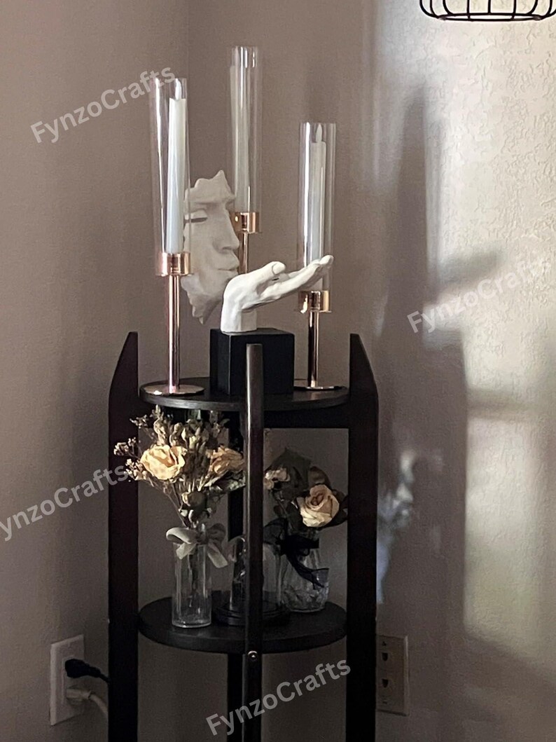 Pu&ograve; includere: Una mensola scura a tre livelli con oggetti decorativi. Presenta tre candelabri in vetro con candele bianche, una scultura bianca di un volto e una scultura di una mano bianca. I ripiani contengono fiori secchi in vasi di vetro.