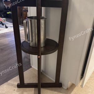 Pu&ograve; includere: Un supporto per distributore di bevande in legno marrone scuro con due ripiani. Una brocca in acciaio inossidabile &egrave; posizionata sul ripiano inferiore. Il ripiano superiore contiene un rubinetto argentato. Il supporto si trova in una stanza con pavimento chiaro.