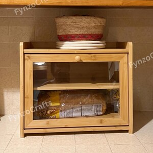 May include: A bamboo bread box with a clear glass door. Inside, a loaf of Nature's butterbread is visible. A woven basket with red and white accents sits on top of the box. The bread box has a wooden knob.