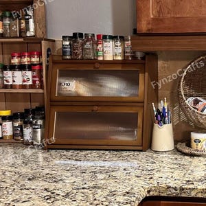May include: A wooden spice rack with two glass-fronted drawers and a top shelf, filled with various spice jars. Additional spice racks and jars are visible on the left. The countertop is granite, and the background includes a wooden cabinet.