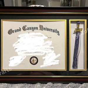 May include: A framed diploma from Grand Canyon University, featuring a gold-trimmed document and a tassel. The frame is dark brown with a black and gold inner border. The tassel is purple and white with a gold 2023 charm.