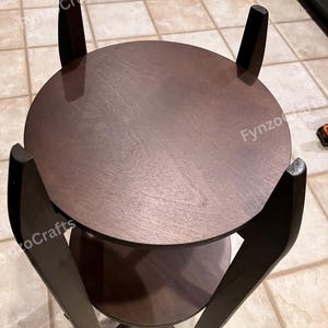 Pu&ograve; includere: Un tavolino in legno marrone scuro a due livelli con piano rotondo e ripiano inferiore. Il tavolo ha quattro gambe angolate e una finitura liscia e lucida. Il tavolo ha un diametro di circa 51 cm.