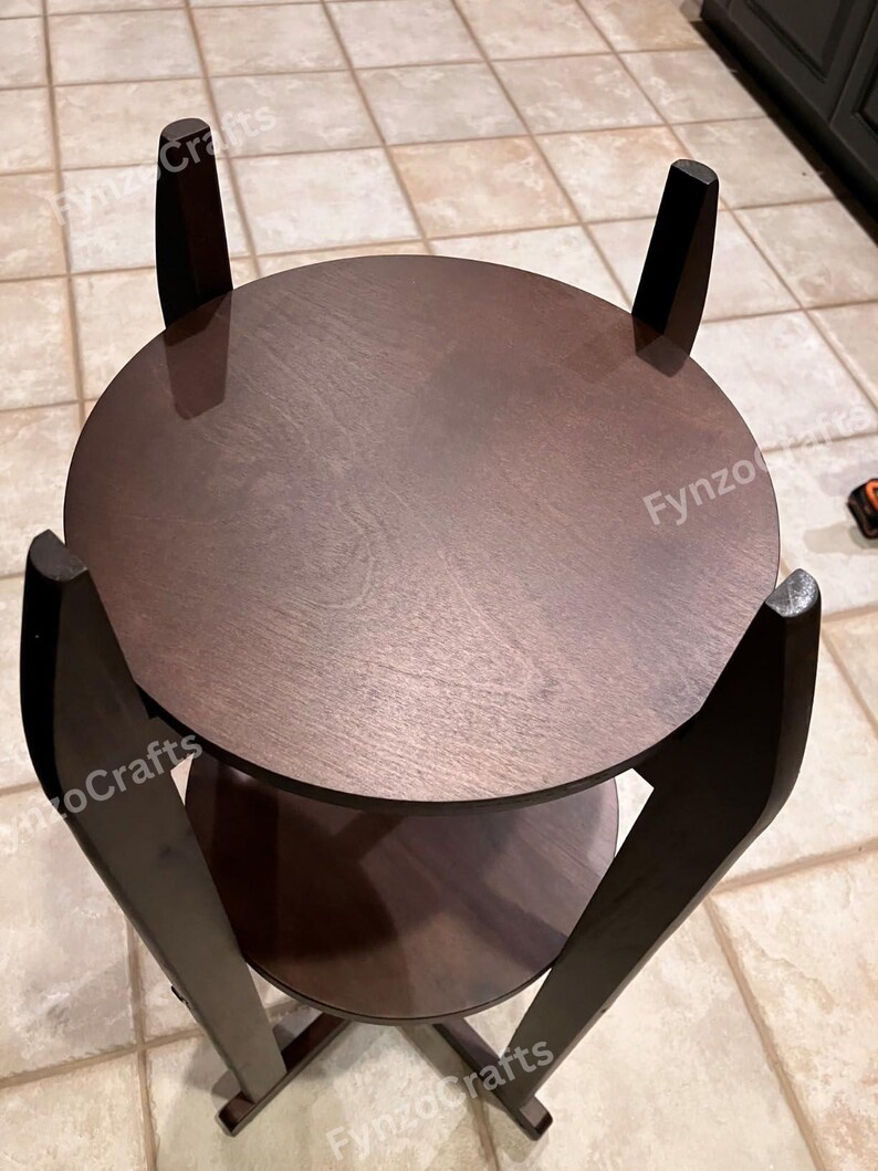 Pu&ograve; includere: Un tavolino in legno marrone scuro a due livelli con piano rotondo e ripiano inferiore. Il tavolo ha quattro gambe angolate e una finitura liscia e lucida. Il tavolo ha un diametro di circa 51 cm.