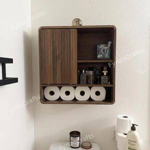May include: A dark brown wooden bathroom storage cabinet with a sliding door, open shelves, and a lower shelf for toilet paper rolls. The cabinet is mounted on a white wall and contains toiletries and accessories.