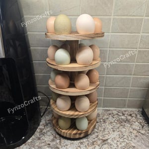 May include: A wooden egg holder with multiple tiers, each holding several eggs of varying colors. The eggs range from white to light brown and green. The holder is made of light-colored wood and has a rustic appearance.