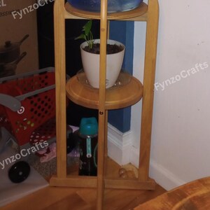 Pu&ograve; includere: Un supporto in legno a tre livelli. Un grande contenitore d'acqua &egrave; posto in cima, una pianta in vaso al centro e una bottiglia d'acqua in basso. Il supporto &egrave; in legno chiaro e ha un design semplice. Il supporto &egrave; in un angolo.