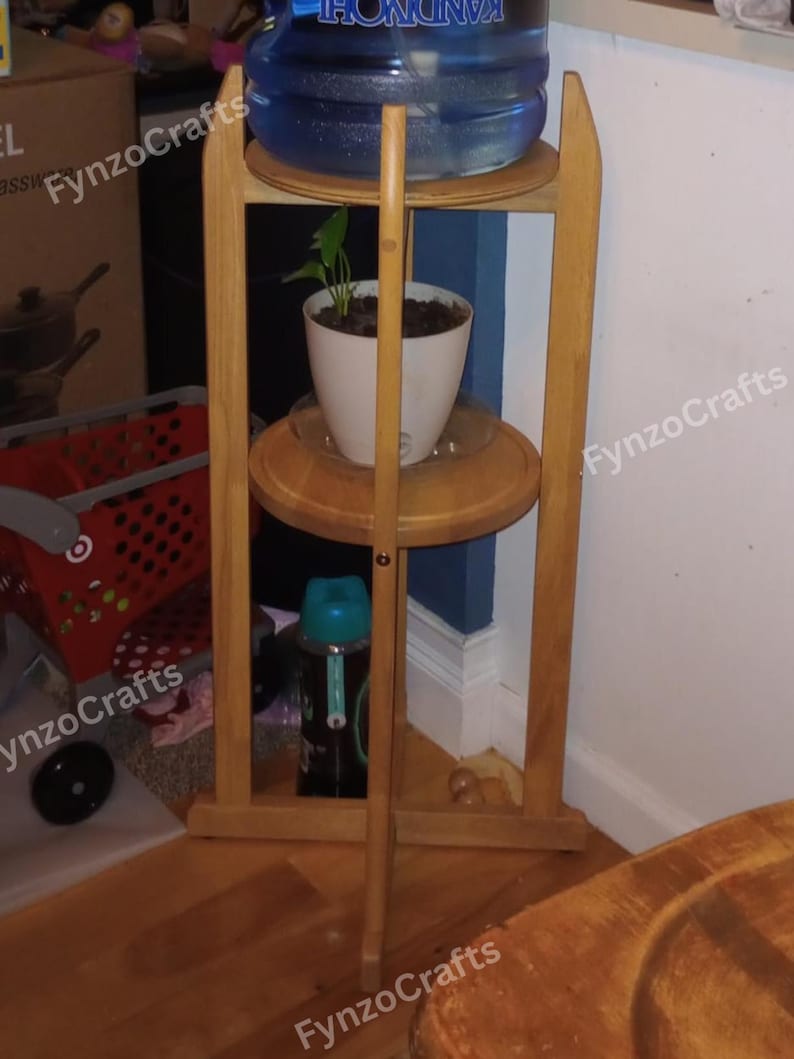 Pu&ograve; includere: Un supporto in legno a tre livelli. Un grande contenitore d'acqua &egrave; posto in cima, una pianta in vaso al centro e una bottiglia d'acqua in basso. Il supporto &egrave; in legno chiaro e ha un design semplice. Il supporto &egrave; in un angolo.