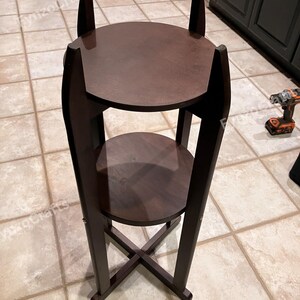 Pu&ograve; includere: Un portapiante in legno marrone scuro con due ripiani circolari. Il supporto ha un design alto e sottile con una base a forma di X per la stabilit&agrave;. La parte superiore del supporto ha due estensioni verticali.