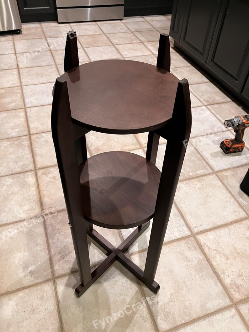 Pu&ograve; includere: Un portapiante in legno marrone scuro con due ripiani circolari. Il supporto ha un design alto e sottile con una base a forma di X per la stabilit&agrave;. La parte superiore del supporto ha due estensioni verticali.