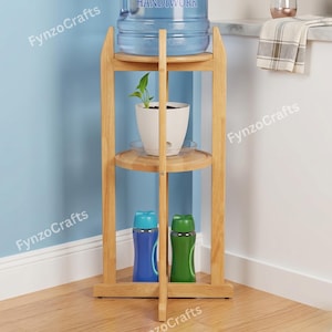 Pu&ograve; includere: Un supporto in legno per refrigeratore d'acqua con tre ripiani. Il ripiano superiore contiene una grande brocca d'acqua. Il ripiano centrale ha una pianta in vaso bianca. Il ripiano inferiore mostra una bottiglia d'acqua blu e verde. Il supporto &egrave; in legno chiaro.
