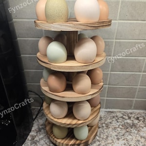 May include: A wooden egg holder with multiple tiers, filled with a variety of eggs in shades of white, tan, and green. The holder has a natural wood finish and the text "FynzoCrafts" is visible on the side.