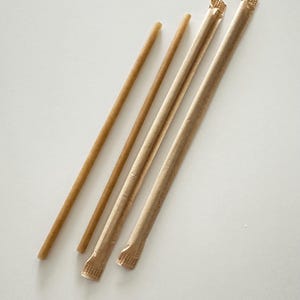 Op de afbeelding: Vier bruine papieren rietjes, twee onverpakt en twee verpakt in papieren hoezen, zijn diagonaal geplaatst tegen een witte achtergrond. De onverpakte rietjes zijn lichter van kleur dan de verpakte. De papieren hoezen hebben een textuur.