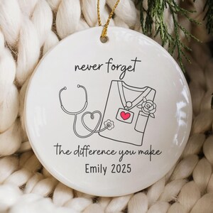 May include: White ceramic ornament with a gold string, featuring a black and red illustration of a stethoscope and medical scrubs. Text reads "never forget the difference you make" and "Emily 2025".