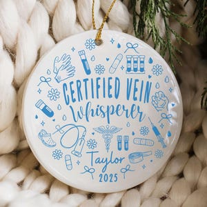 May include: A white ceramic ornament with blue medical-themed illustrations and text. The ornament says "CERTIFIED VEIN Whisperer Taylor 2025". It is hanging by a gold string, set against a white knitted background and evergreen branches.