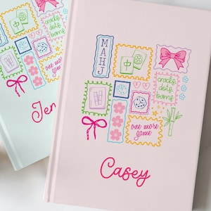 May include: Two hardcover journals in light blue and light pink, each featuring a colorful graphic print of Mahjong tiles, dice, bows, and the text "MAHJ", "cracks dots bams", and "one more game". The journals are personalized with the names Jen and Casey.