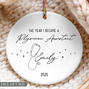 May include: A round white ceramic ornament featuring black text that reads THE YEAR I BECAME A Physician Assistant, the name Emily, and the year 2026. A black line drawing of a stethoscope and small star icons are centered on the ornament surface.