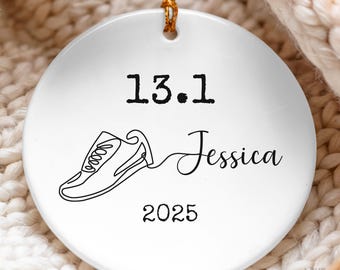 May include: A round white ceramic ornament featuring a black line art drawing of a running shoe, the text 13.1, the name Jessica in script, and the year 2025. The ornament hangs from a gold string and has a smooth, glossy finish.