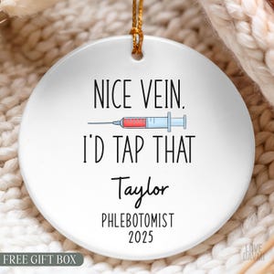 May include: A round white ceramic ornament featuring a graphic of a syringe with red liquid. The text reads: NICE VEIN. I'D TAP THAT, Taylor, PHLEBOTOMIST, 2025. The ornament has a small hole at the top with a gold string for hanging purposes.