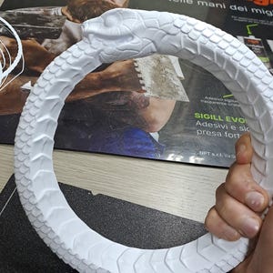 May include: A white circular sculpture depicting a snake biting its own tail, known as an Ouroboros. The object features detailed, raised scale textures covering the entire surface of the serpent's body, with a clearly defined head and eye.