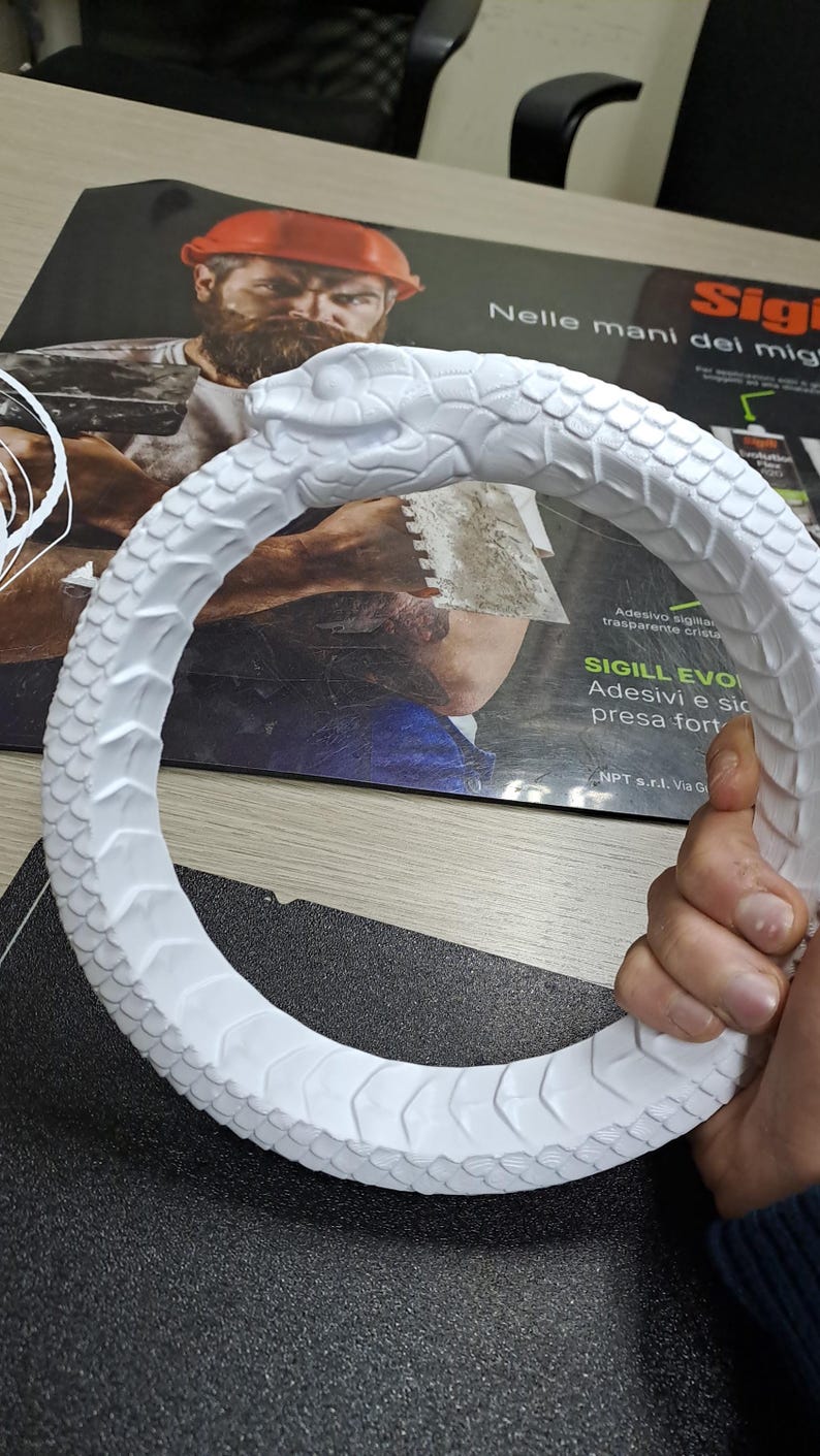 May include: A white circular sculpture depicting a snake biting its own tail, known as an Ouroboros. The object features detailed, raised scale textures covering the entire surface of the serpent's body, with a clearly defined head and eye.