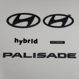 May include: A set of matte black automotive emblems including two Hyundai oval logos, the word hybrid in lowercase, the text HTRAC in a rectangular border, and the word PALISADE in uppercase letters, all featuring a clean, flat finish.