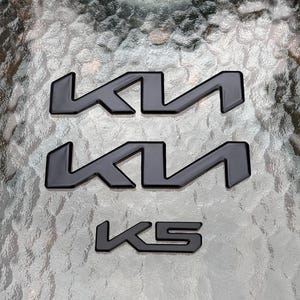 May include: Three black car emblems. Two feature the stylized letters "K" and "I" in a connected design. The third emblem reads "K5" in a similar font. All are set against a textured, reflective surface.