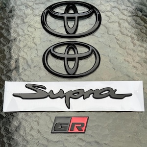 May include: A collection of black car emblems and decals. The image includes two Toyota emblems, a "Supra" decal, and a "GR" emblem with a red background. The items are displayed on a reflective surface.