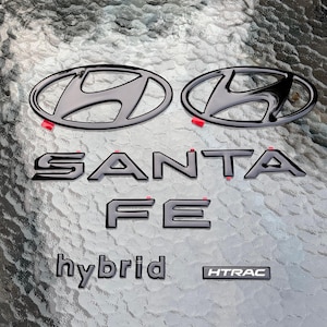 May include: A set of glossy black automotive emblems including two Hyundai logos, the text SANTA FE, the word hybrid, and a rectangular HTRAC badge. Each piece features a smooth finish and small red adhesive tabs visible on the edges for installation.