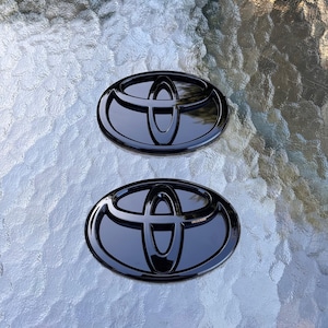 Puede incluir: Dos insignias de emblema Toyota negras brillantes. Cada insignia tiene forma ovalada con el logotipo de Toyota en el centro. Las insignias se muestran sobre una superficie texturizada de color claro.