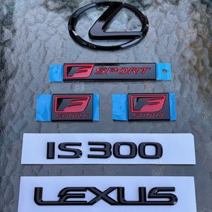 Peut inclure: Ensemble d'emblèmes et d'insignes de voiture Lexus noirs et rouges. Comprend un logo Lexus, des badges "F SPORT" et les lettres "IS 300" et "LEXUS". Les emblèmes sont sur un support blanc.