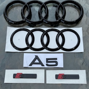 May include: Black car emblem set featuring the Audi rings logo, an "A5" emblem, and two "S line" badges with red accents. The emblems are displayed on a light-colored surface.