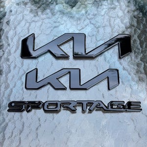 Könnte beinhalten: Drei schwarze Kia-Embleme und ein Sportage-Emblem werden auf einer strukturierten, hellfarbigen Oberfläche angezeigt. Zwei Kia-Embleme sind gestapelt, und das Sportage-Emblem befindet sich darunter. Die Kia-Embleme haben ein stilisiertes, eckiges Design.