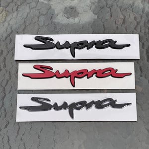 以下が含まれることがあります： 3つのSupraエンブレムデカール。2つは黒、1つは赤です。各デカールは、白い長方形の背景に、様式化されたフォントで「Supra」という単語を表示しています。デカールは、テクスチャ加工された表面に表示されます。