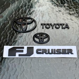 May include: Black Toyota emblems and FJ Cruiser lettering. The Toyota logo is displayed twice, with the word "TOYOTA" in block letters. The FJ Cruiser lettering is on a white backing.